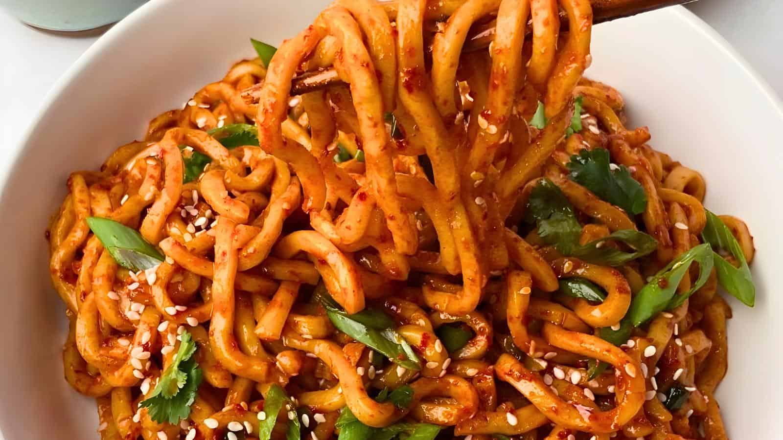 A bowl of spicy noodles coated in a red sauce, garnished with sesame seeds, green onions, and cilantro. A pair of chopsticks lifts a portion of the noodles from the bowl.