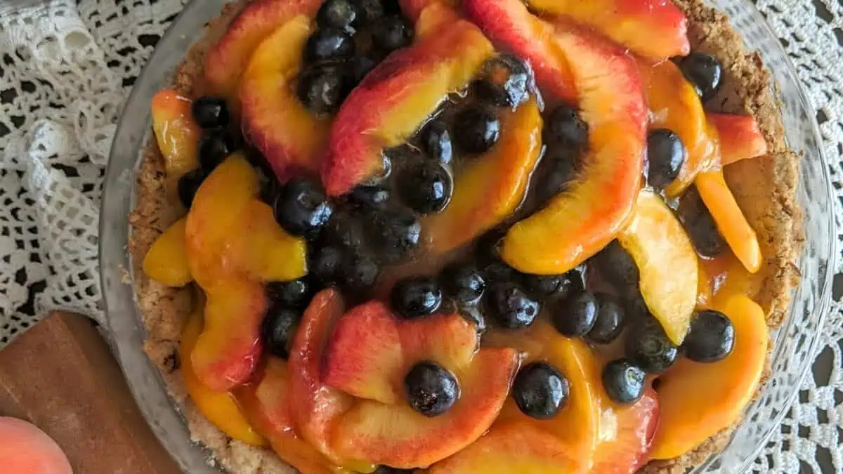A delectable fruit tart with a flaky crust is topped with succulent sliced peaches and plump blueberries, displayed on a lace tablecloth. The tart's glistening glaze enhances the vibrant colors of the fresh fruits.