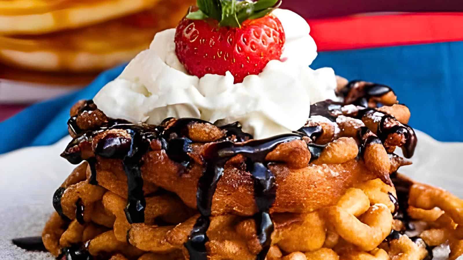 A funnel cake topped with whipped cream, chocolate syrup, and a strawberry. Blurred in the background is a stack of pancakes.