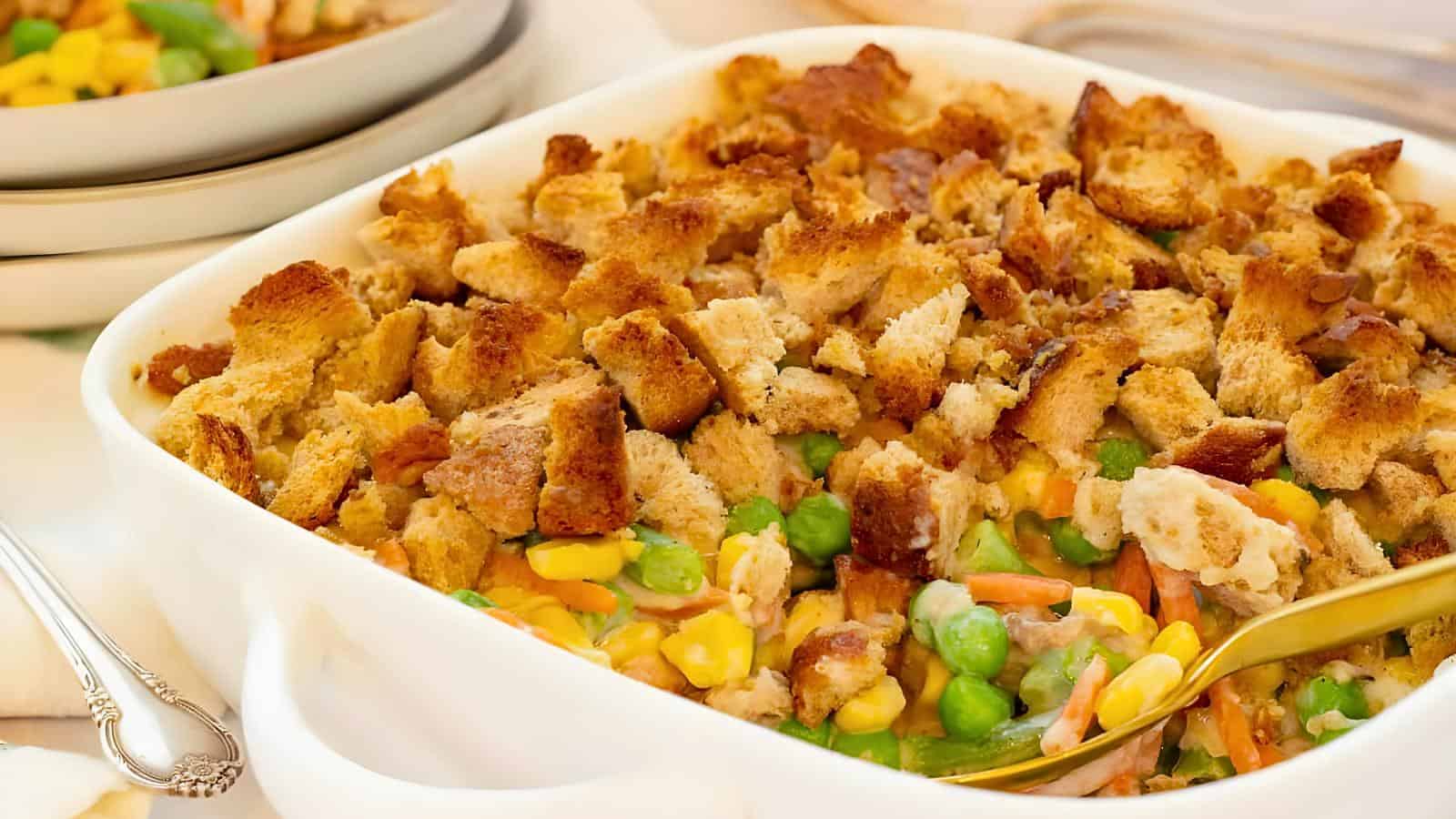 A white ceramic container filled with mixed vegetables and topped with toasted bread pieces, with a serving spoon placed inside.