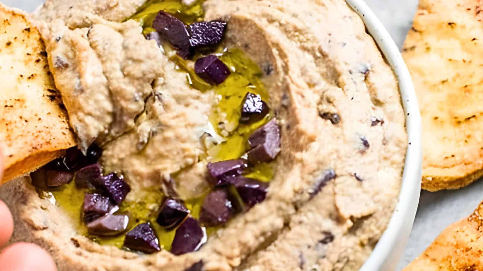 A bowl of hummus topped with olive pieces and drizzled with olive oil, accompanied by pita bread slices.