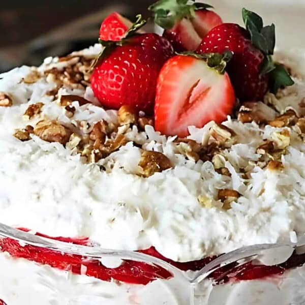 A layered dessert in a glass bowl with whipped cream, strawberries, nuts, and coconut flakes, topped with fresh strawberries.