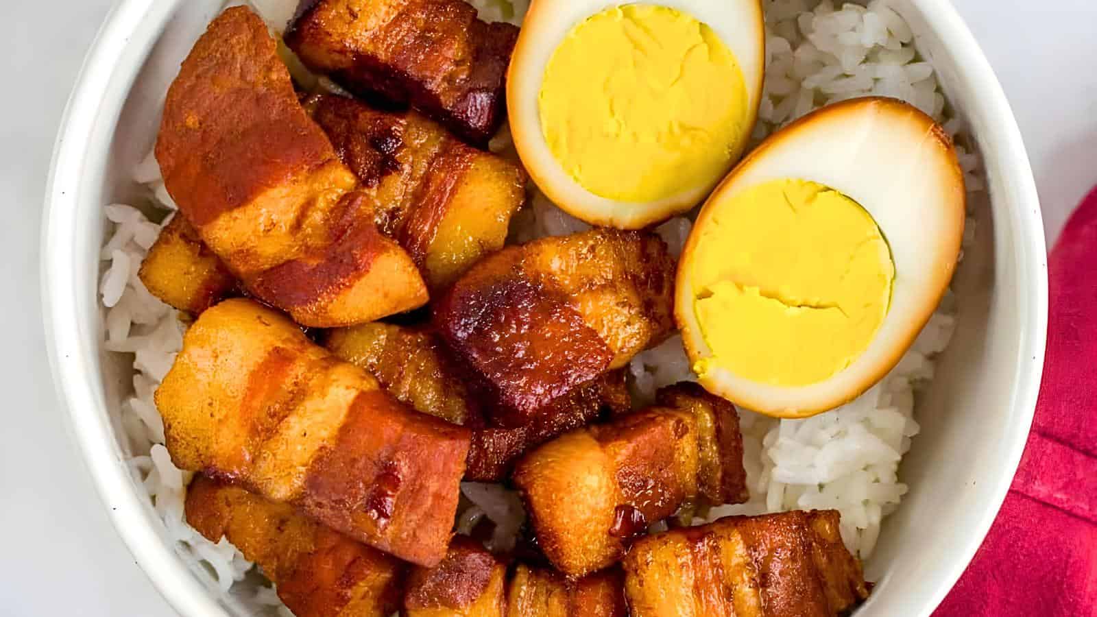 A bowl of rice topped with slices of caramelized pork belly and two halves of a soft-boiled egg.