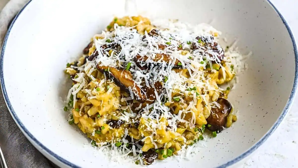 A bowl of creamy mushroom risotto topped with grated Parmesan cheese and garnished with chopped green herbs. The dish features a golden-brown color with visible pieces of mushrooms mixed in. The bowl is white with a blue rim, placed on a light-colored surface.