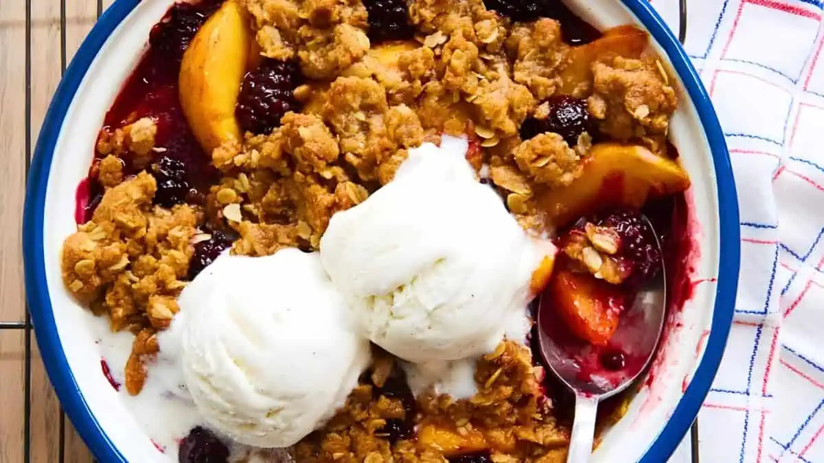 A close-up of a fruit crisp dessert in a bowl, topped with two scoops of vanilla ice cream. The crisp includes a variety of fruits and a crumbly oat topping. A spoon is also visible in the bowl, and a checkered napkin is placed beside it.