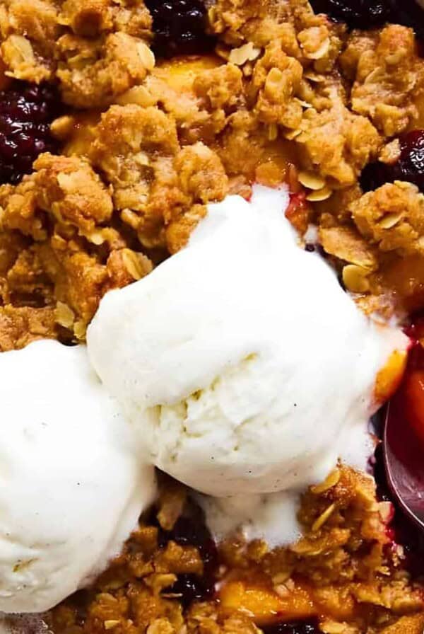 A bowl of fruit crumble topped with two scoops of vanilla ice cream, featuring a mix of baked fruits and an oat crumble, with a spoon partially dipped into the crumble. Texture of ice cream and crumble visible.