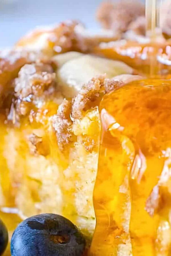 A slice of cake garnished with blueberries and crumbled pieces, being drizzled with honey.