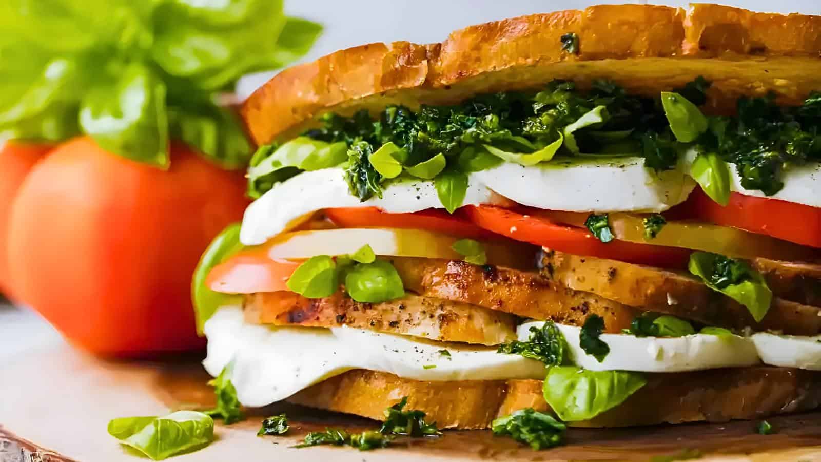 Thick slices of bread filled with tomato, mozzarella, basil leaves, and pesto. Background includes tomatoes.