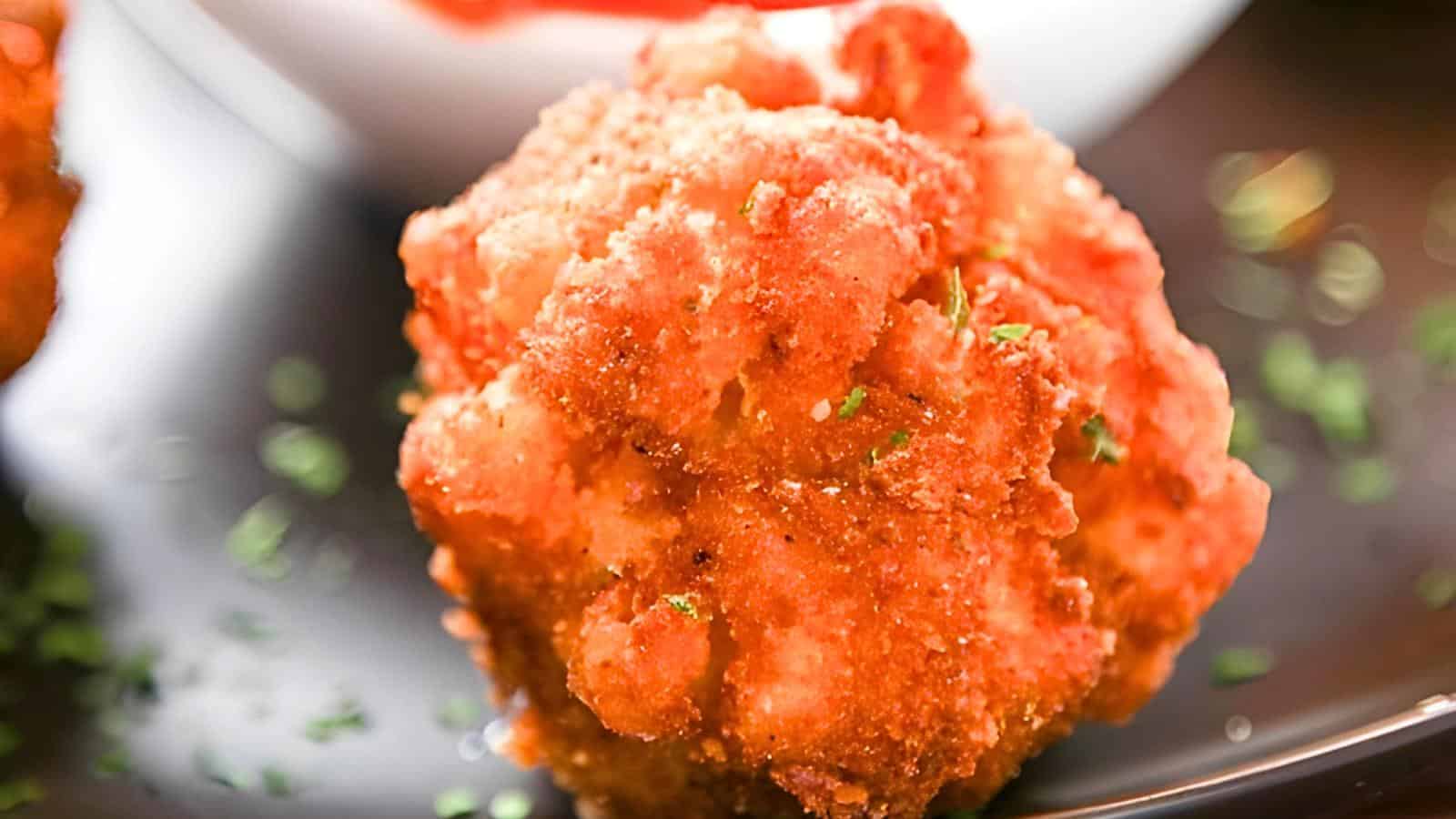 A golden, crispy fried mac and cheese garnished with herbs on a dark plate, with a hint of red sauce in the background.
