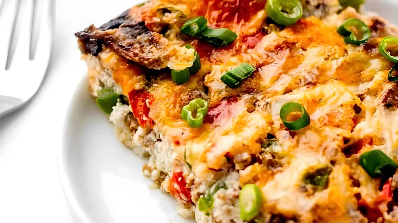 A slice of baked casserole with melted cheese, green peppers, tomatoes, and green onions on a white plate next to a metal fork.