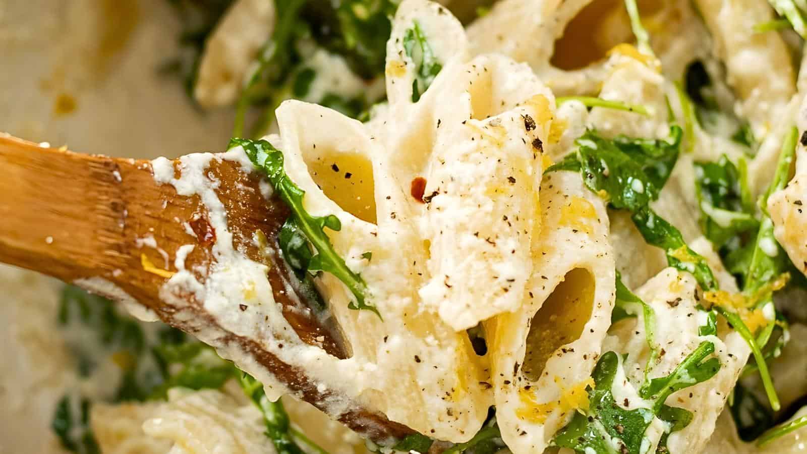 Pasta coated in white sauce with spinach, garnished with black pepper on a wooden spoon.