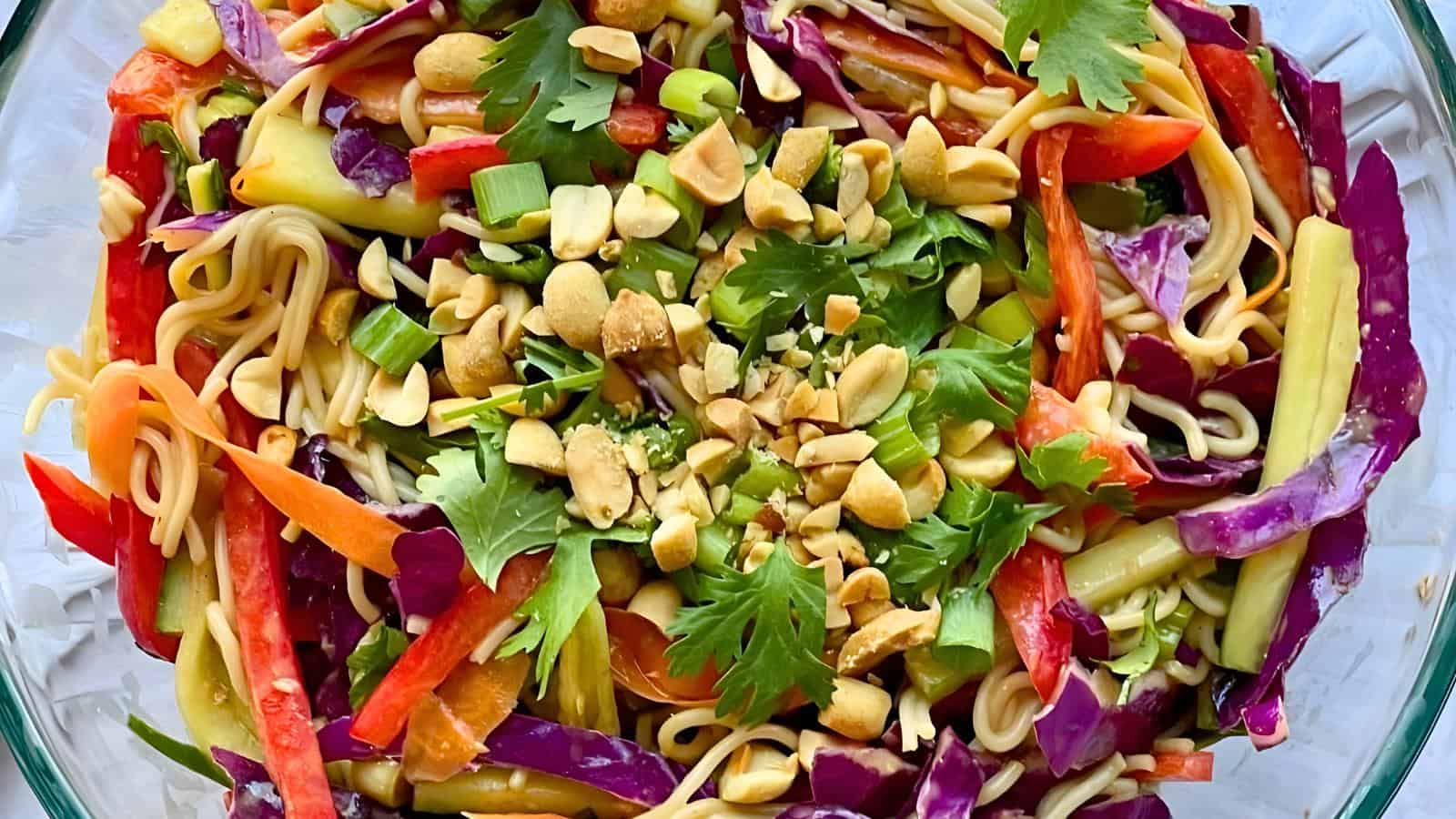 A vibrant salad with noodles, red cabbage, sliced red and yellow peppers, green onions, and cilantro, garnished with chopped peanuts in a glass bowl.