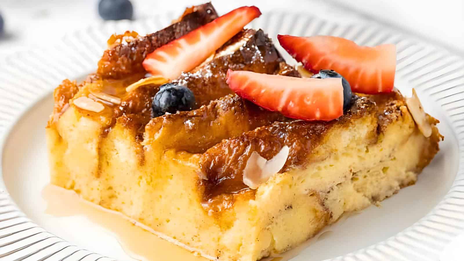 A slice of French toast topped with caramelized sugar, sliced strawberries, blueberries, almond slivers, and syrup on a white plate.