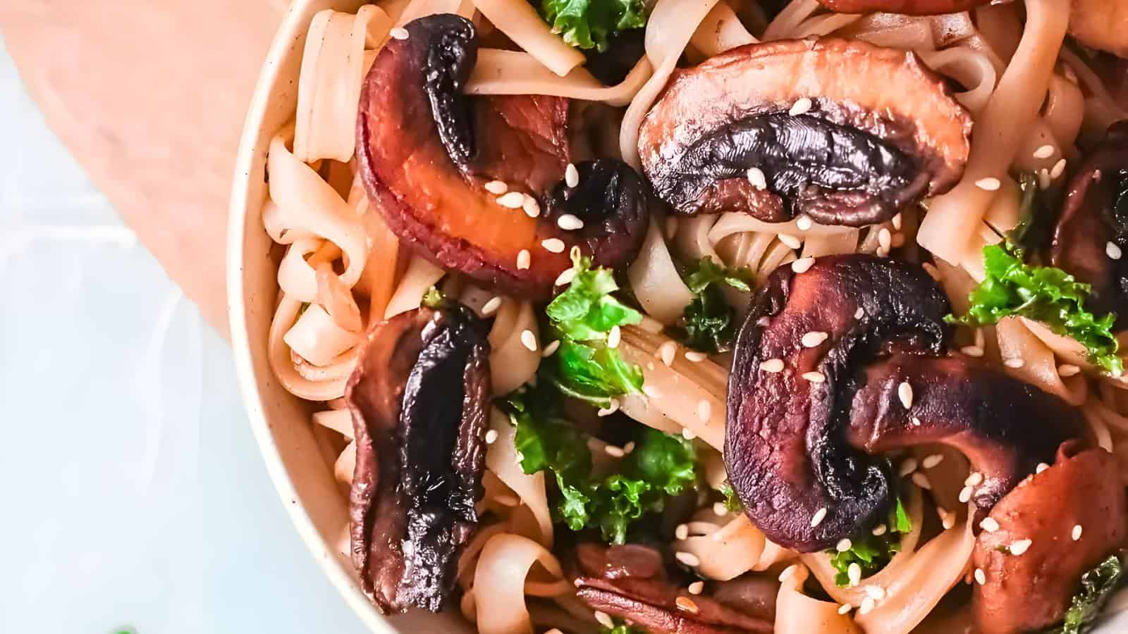 A close-up of a bowl of pasta with sautéed mushrooms, vibrant green kale, and sesame seeds. The noodles are intertwined with the earthy mushrooms and sprinkled with seeds, creating a visually appealing and appetizing dish.