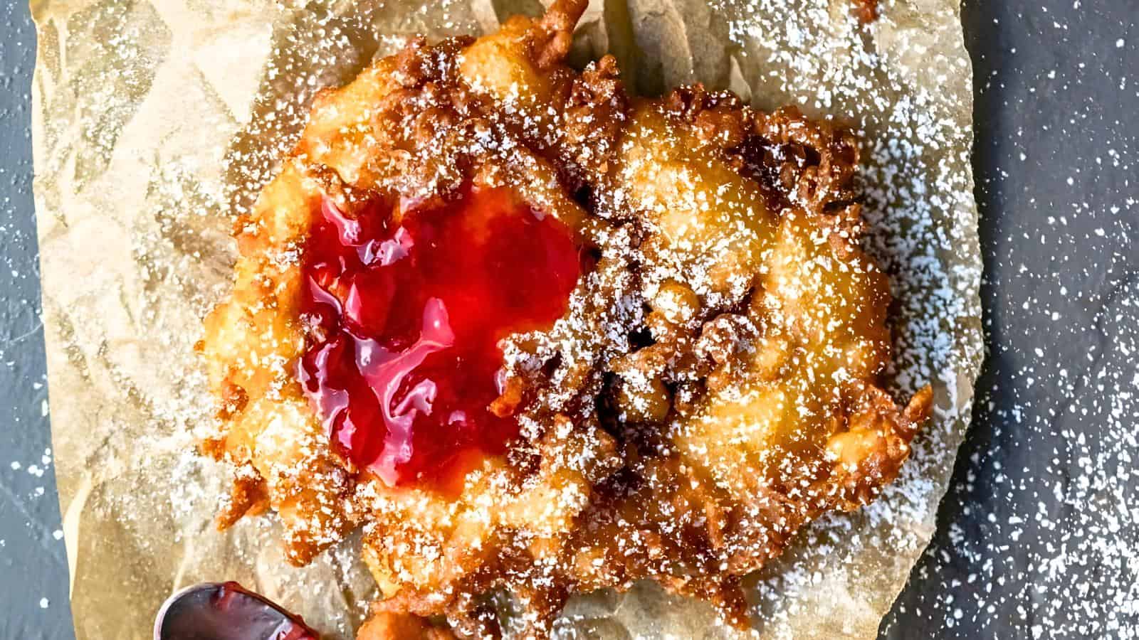 A fried pastry dusted with powdered sugar, topped with bright red jelly filling, placed on parchment paper.