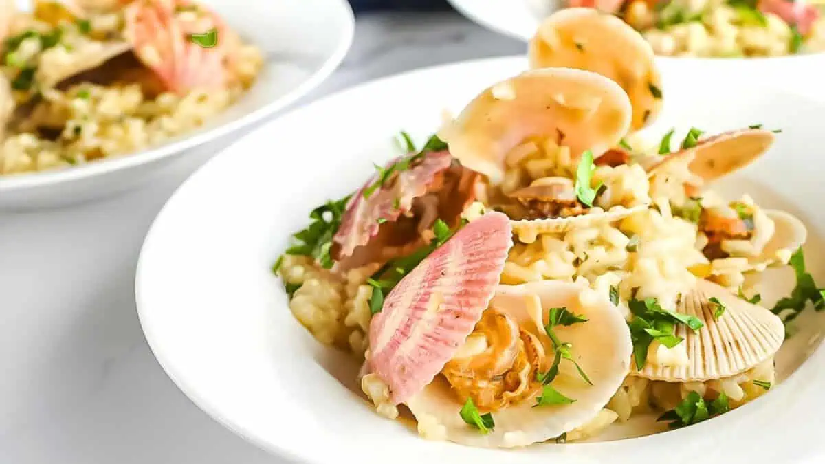 A white bowl filled with creamy risotto, garnished with several pink and white clams, and fresh parsley. The dish looks elegantly plated and sits on a white surface. In the background, there are additional bowls of the same dish.
