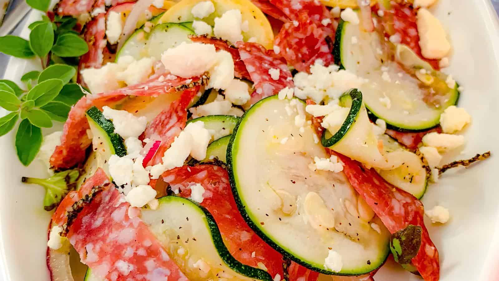 Zucchini slices, salami, yellow squash, and feta cheese garnished with basil on a white dish.