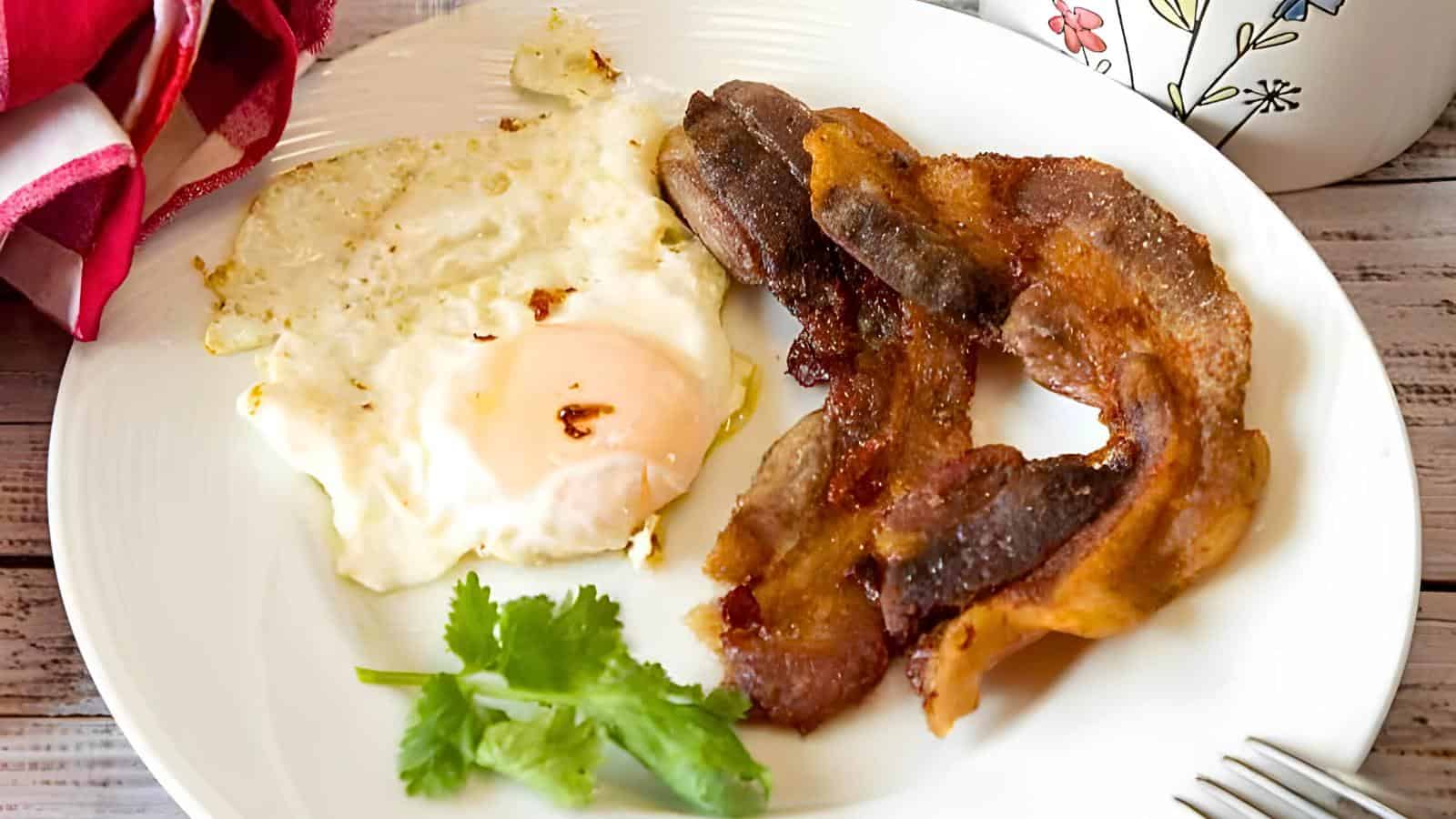 A white plate with a fried egg, a few strips of cooked bacon, and a sprig of cilantro. A mug with a floral design and a red napkin in the background.