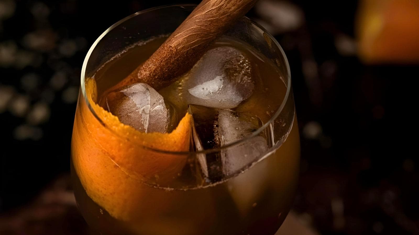 A cocktail garnished with an orange peel and ice cubes, stirred with a wooden muddler.