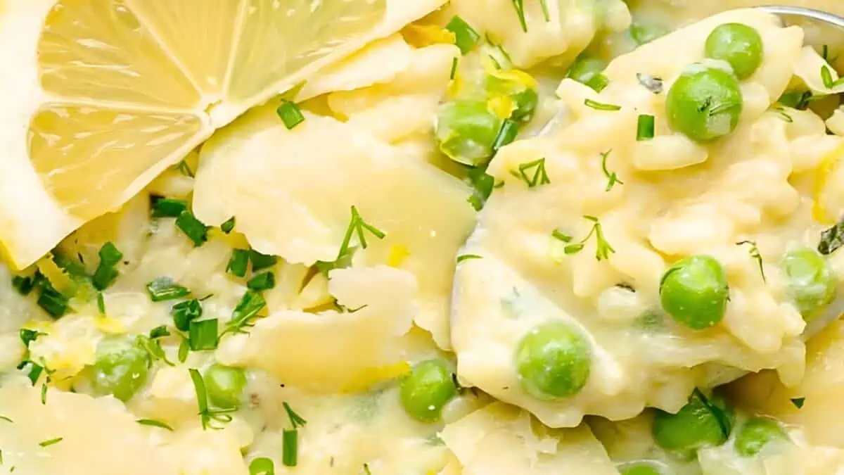 Close-up of a creamy vegetable risotto with green peas, zucchini, and fresh herbs, garnished with lemon slices and chopped chives. The rich, velvety texture is visible, with vibrant green peas and finely diced zucchini adding color to the dish.