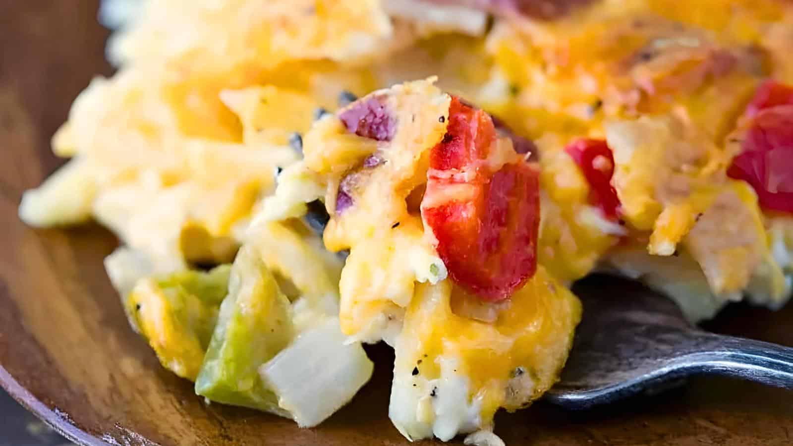 A forkful of cheesy breakfast casserole with tomato pieces, diced green peppers, and onions on a wooden plate.