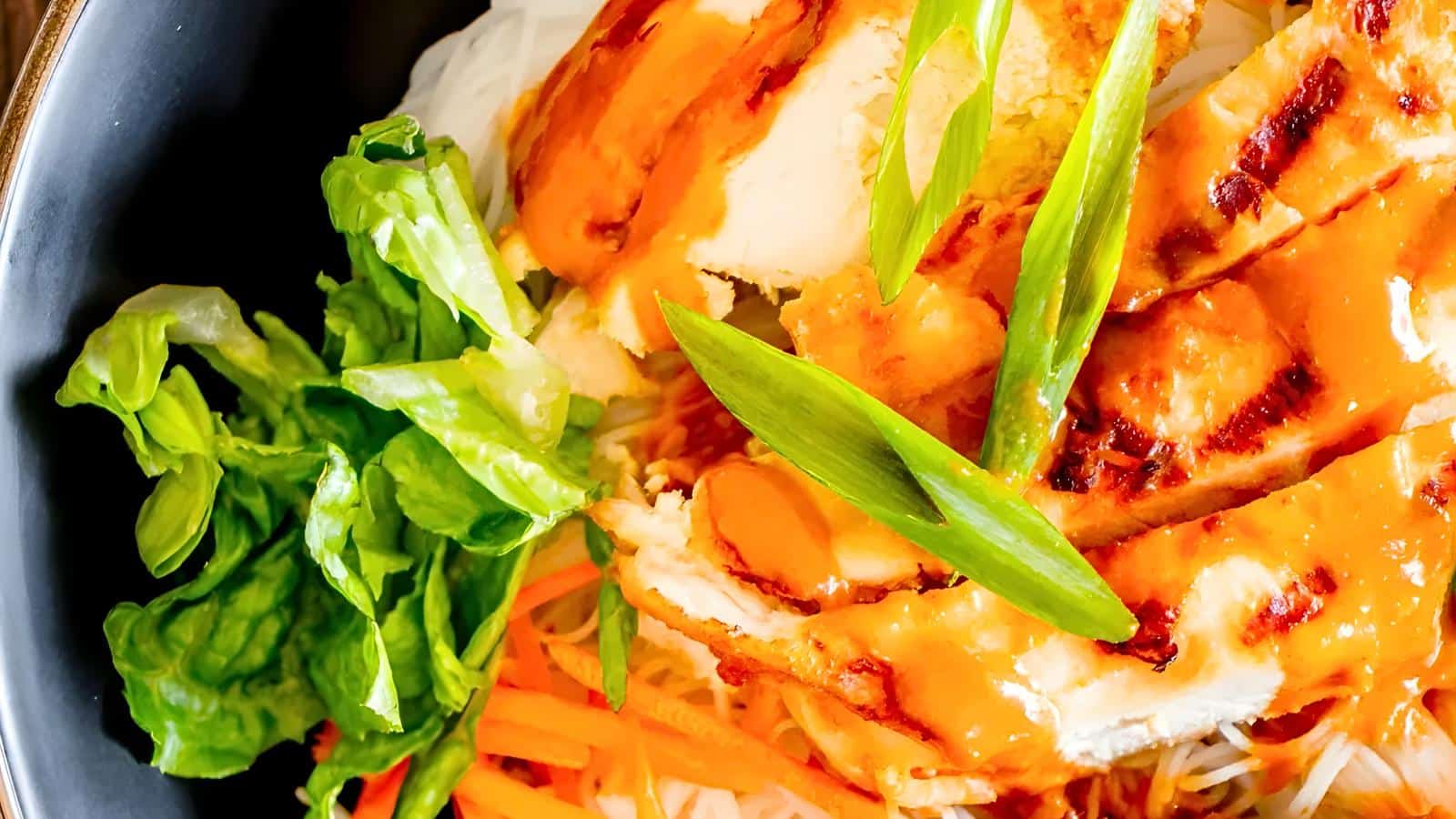 A close-up of a dish featuring grilled chicken slices drizzled with orange sauce, placed on a bed of vermicelli noodles with green lettuce and julienned carrots, garnished with fresh green onions.