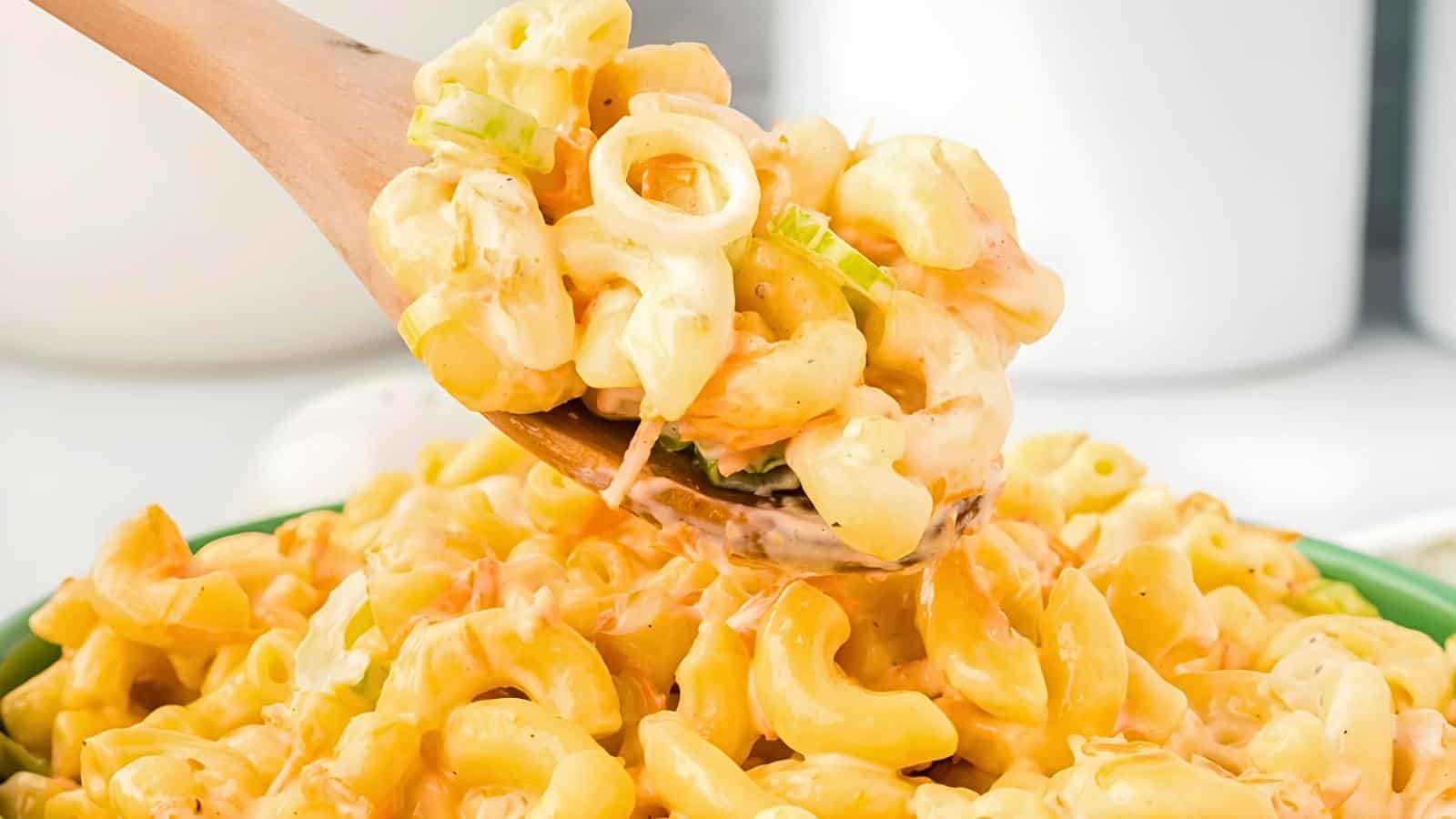 A wooden spoon scooping creamy macaroni and cheese from a green bowl, showing pasta mixed with a cheese sauce and a green garnish.
