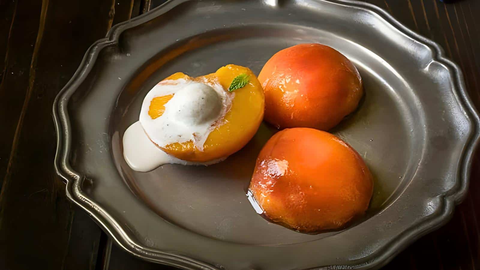 A metal plate with two whole glazed peaches, one half peach topped with a scoop of vanilla ice cream, and a mint leaf garnish.