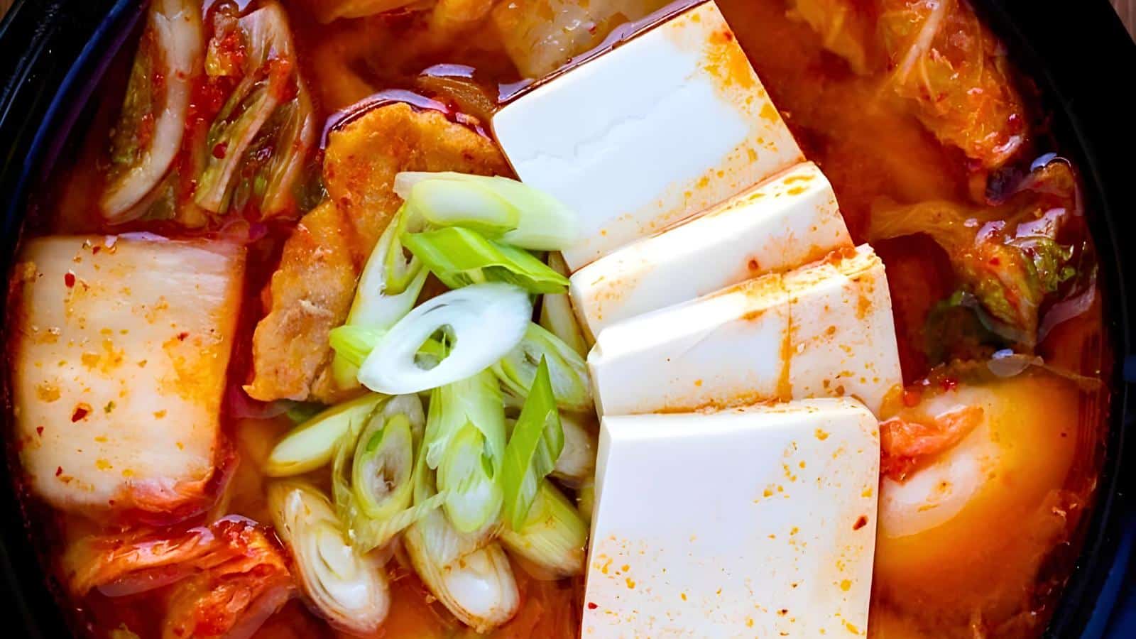 A bowl of kimchi stew with tofu, green onions, cabbage kimchi, and sliced meat in a spicy red broth.