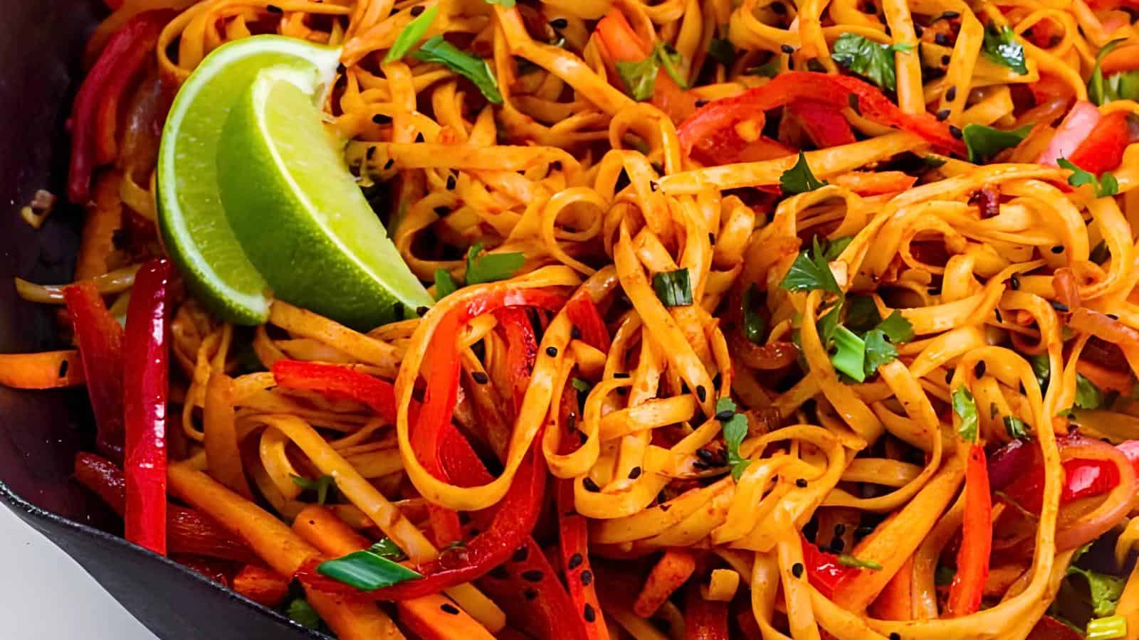 Close-up of a colorful noodle dish with sliced red peppers, carrots, and fresh green herbs. A wedge of lime is placed on top for garnish. The noodles appear tangy and well-seasoned.