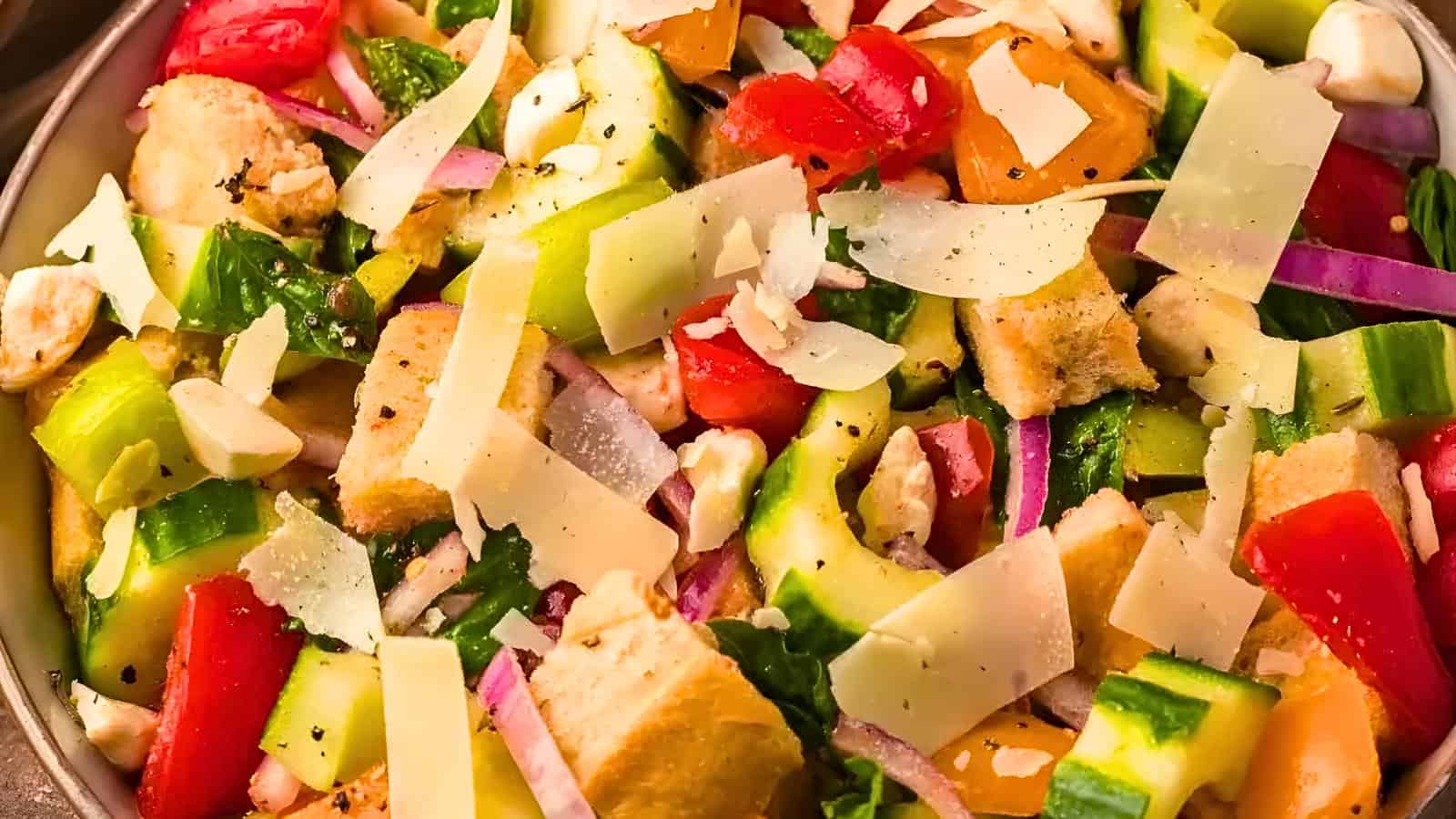 A bowl of chopped cucumbers, tomatoes, red onions, mozzarella balls, croutons, and Parmesan shavings.