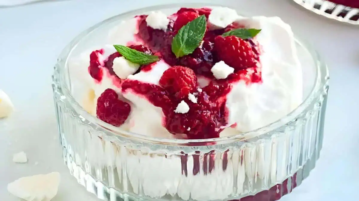A glass dish filled with a dessert made of whipped cream, topped with raspberry sauce, fresh raspberries, and garnished with small mint leaves. Small pieces of meringue are scattered on top of the dessert.