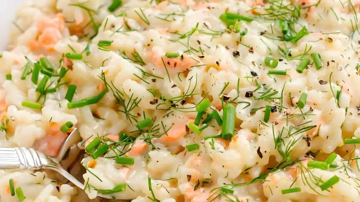 A close-up view of a creamy rice dish garnished with fresh green herbs such as dill and chives. Small pieces of salmon are mixed throughout the dish, and a fork is positioned on the left side, slightly submerged in the creamy mixture.