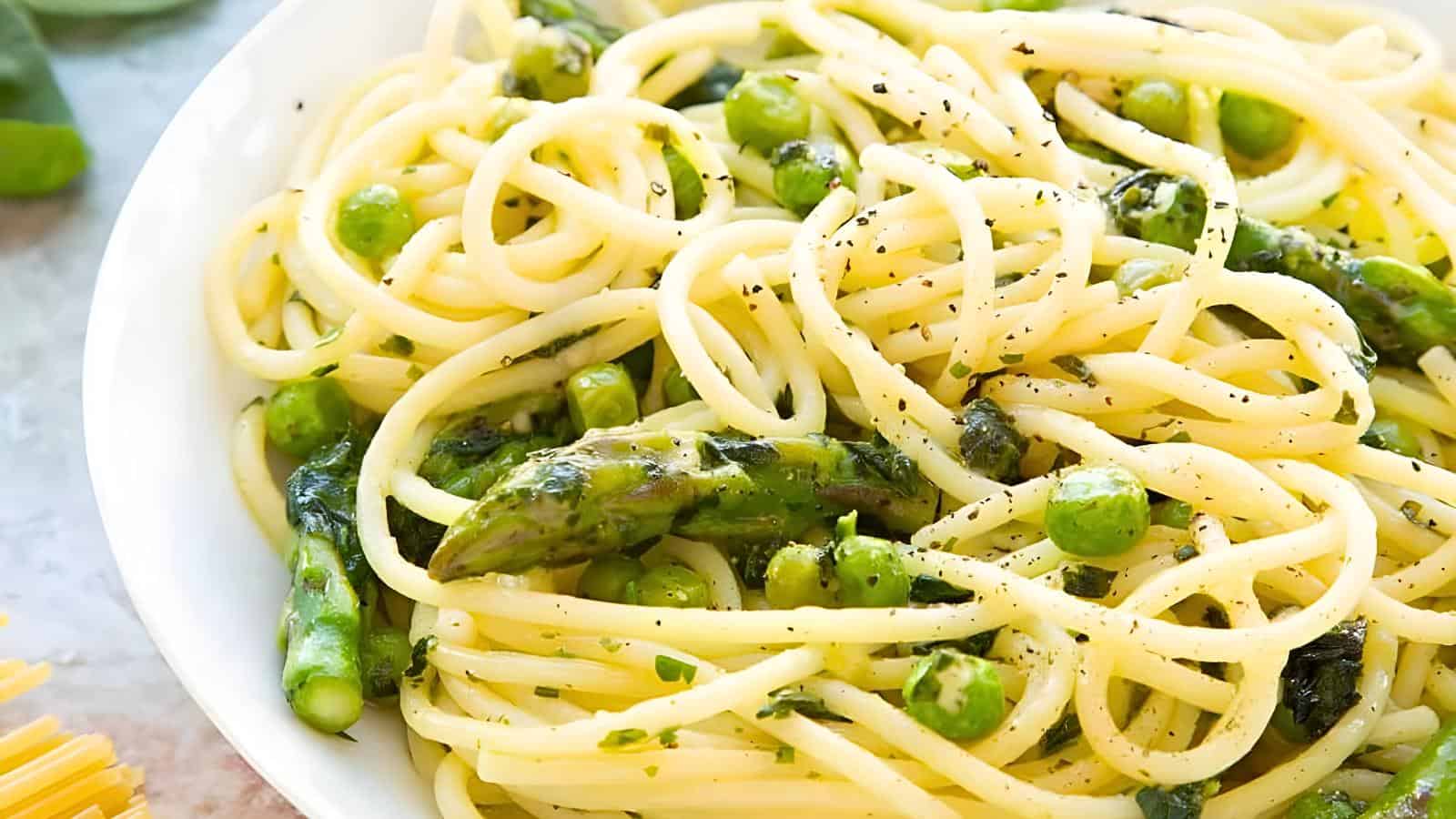 A close-up of a bowl of spaghetti pasta mixed with asparagus, peas, and herbs, sprinkled with black pepper. The dish is vibrant and garnished with freshly ground seasoning.