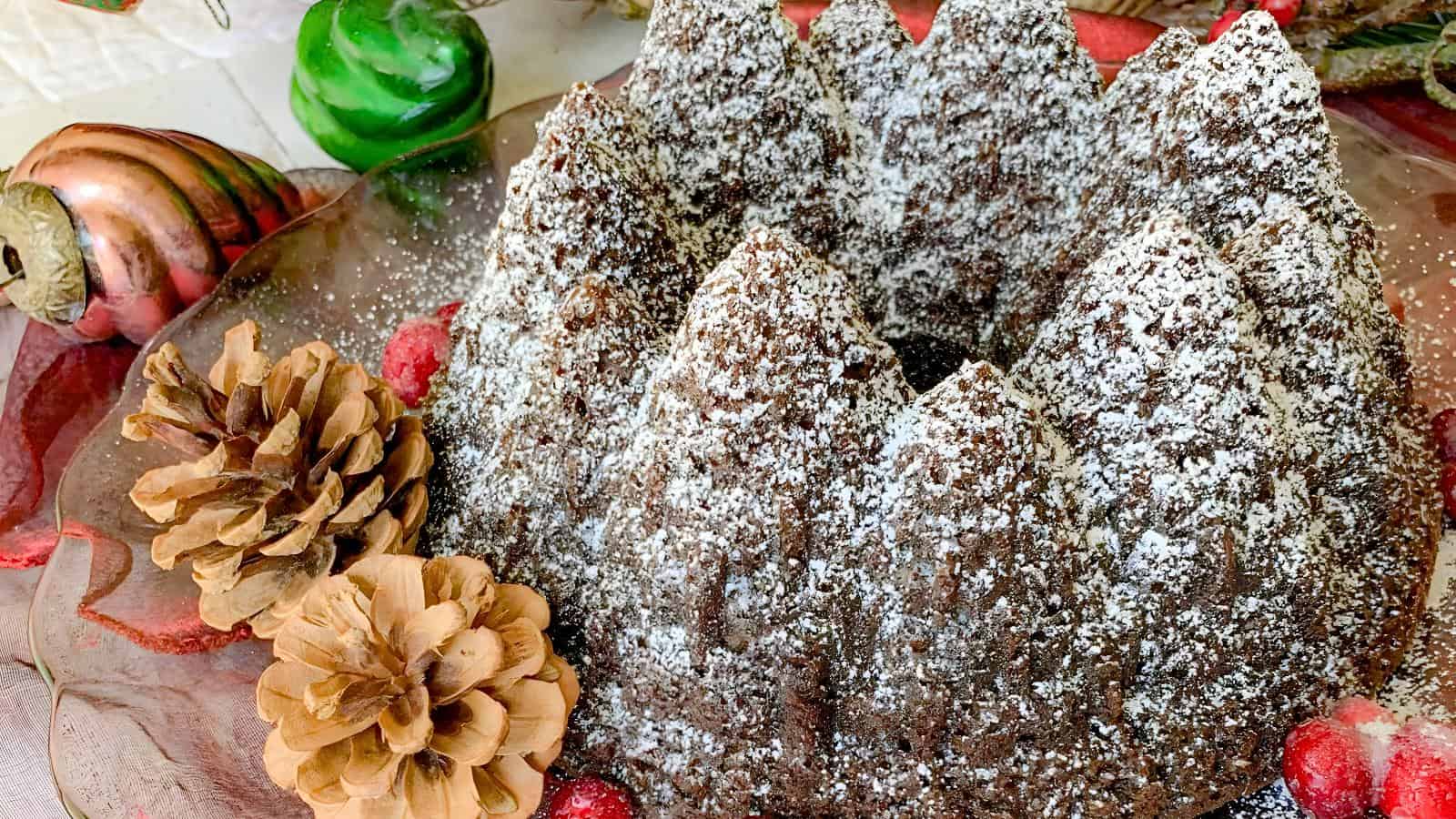 A chocolate bundt cake with powdered sugar sits on a decorative plate. Pine cones and Christmas ornaments are arranged next to the cake.