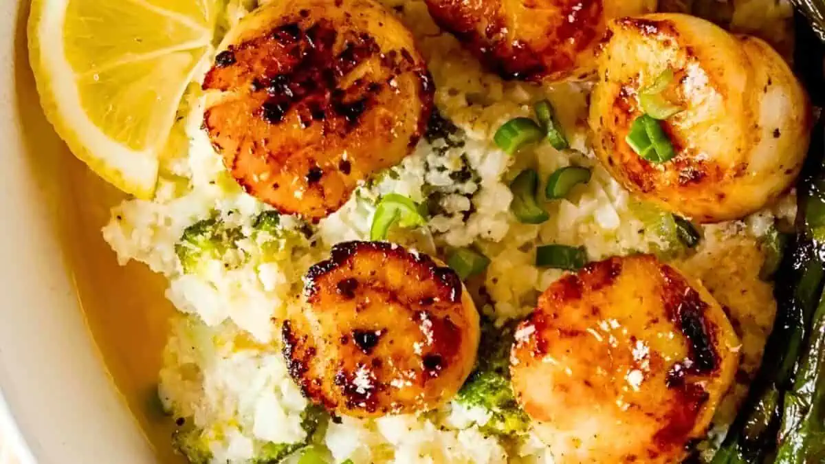A close-up of a dish featuring seared scallops placed on a bed of mashed cauliflower mixed with broccoli. Garnished with chopped green onions and a lemon wedge on the side. The dish appears freshly prepared with a golden-brown finish on the scallops.