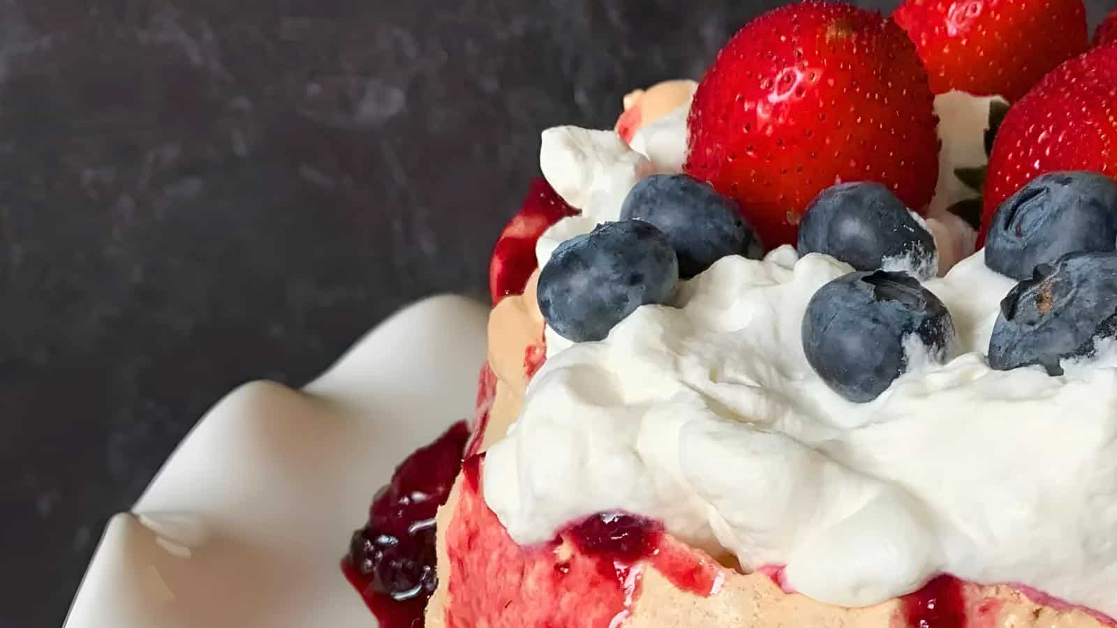 Close-up of a dessert topped with whipped cream, fresh strawberries, and blueberries. The dessert appears to have layers, with hints of red sauce drizzling down the sides. The background is dark, contrasting with the vibrant colors of the fruit.