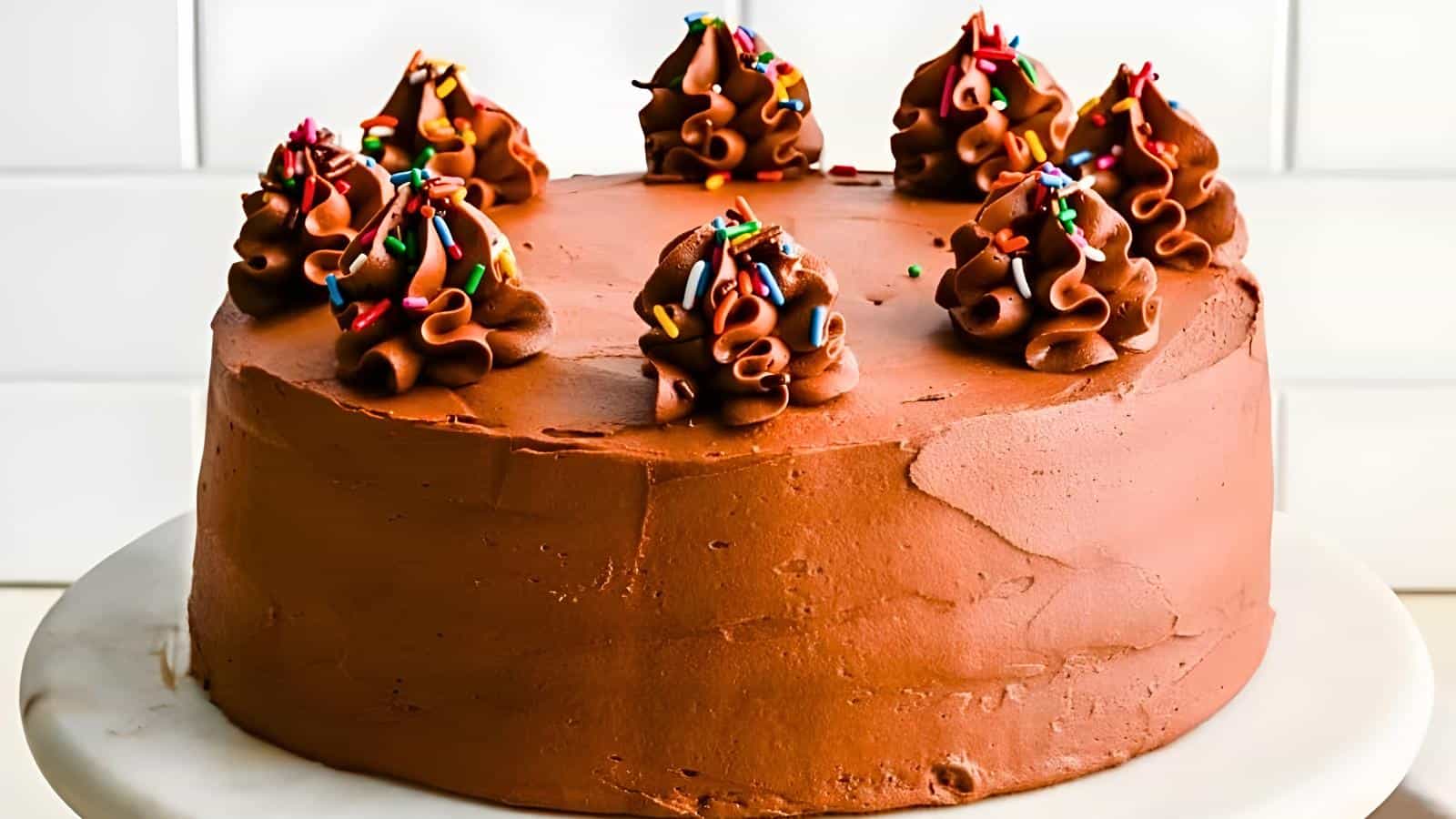 A round chocolate cake on a white cake stand, topped with swirls of chocolate frosting and colorful sprinkles.