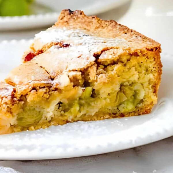 A slice of grape pie on a white plate with a golden brown crust and moist, green grape filling. Small bunch of green grapes on the side.