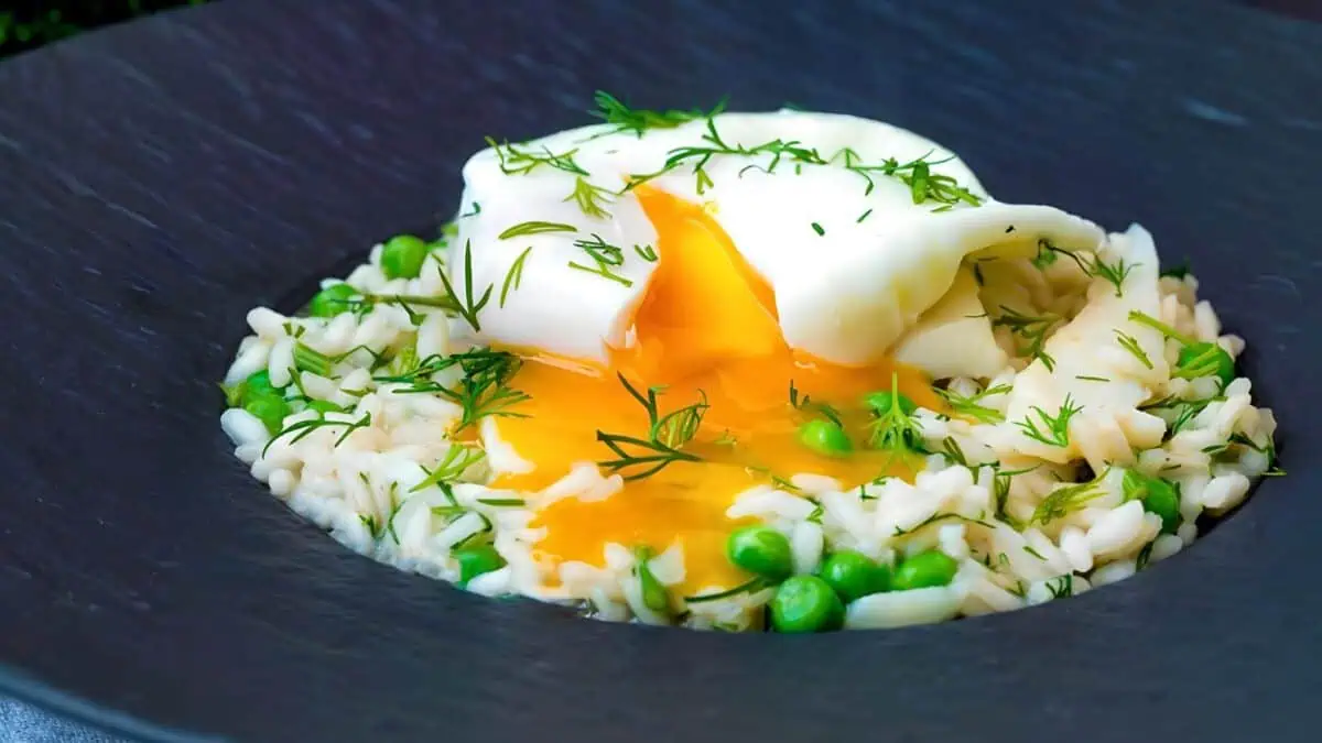 A gourmet dish featuring a bed of white rice mixed with green peas, topped with a poached egg. The egg yolk is slightly broken, releasing a golden ooze over the rice. Fresh dill sprigs are sprinkled over the dish, adding a touch of green. Served on a dark plate.