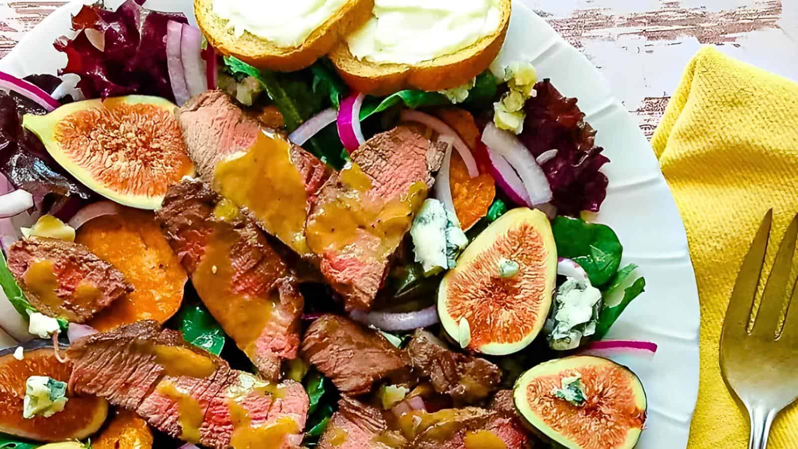 A plate of steak salad with fig slices, red onions, and greens, topped with dressing. Garlic bread on the side. A yellow napkin and a fork next to the plate.