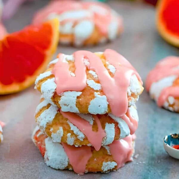 Three cookies with pink icing are stacked beside a halved grapefruit. Additional cookies and grapefruit slices appear in the background. A small spoon is nearby.