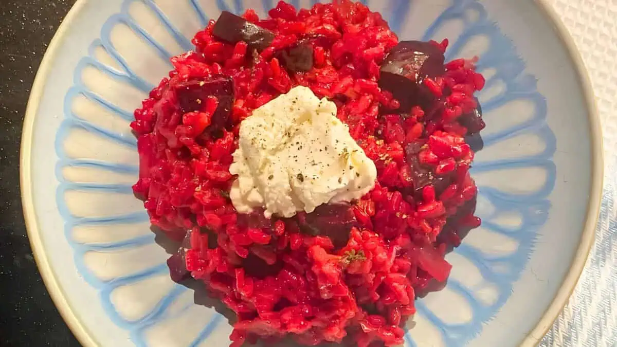 A plate of vibrant red beetroot risotto is topped with a dollop of creamy white cheese and a sprinkle of black pepper. The dish is served on a blue and white patterned plate.