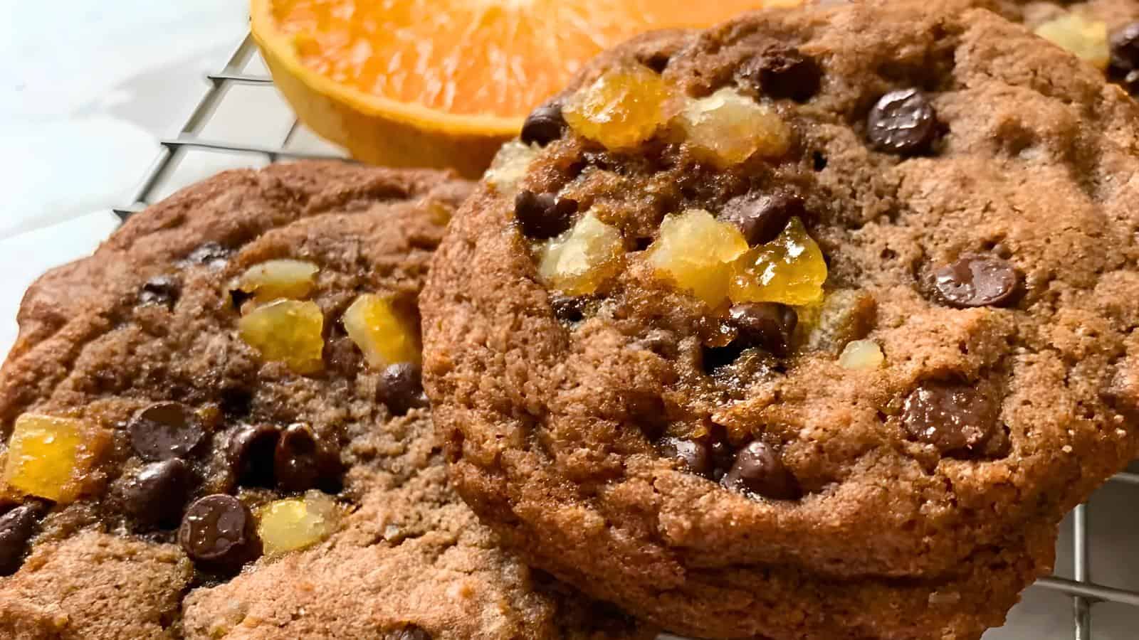 Chocolate chip cookies with small chunks of candied ginger, placed in front of an orange slice on a cooling rack.