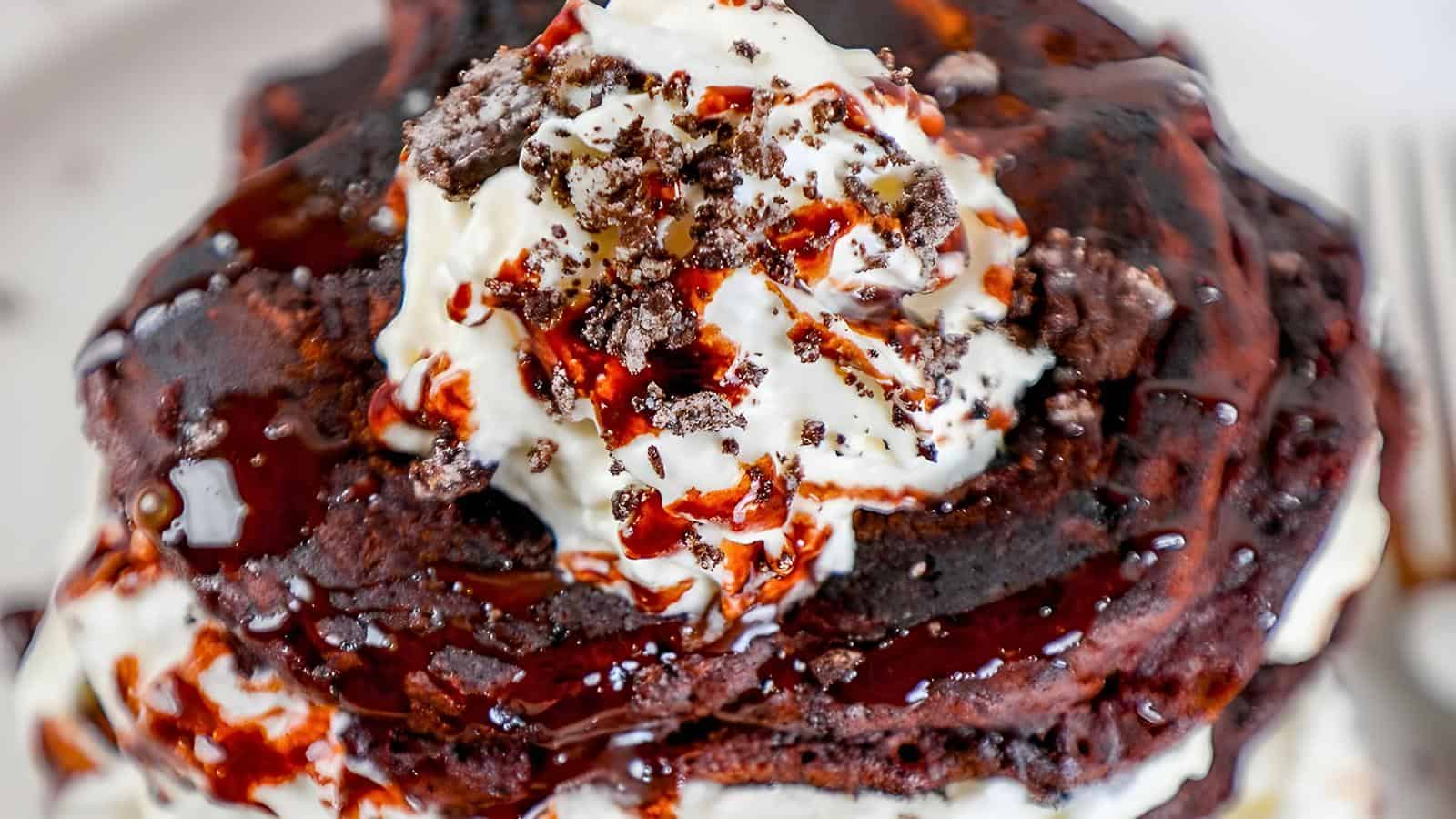 A stack of chocolate pancakes with whipped cream, chocolate drizzle, and crushed cookies.