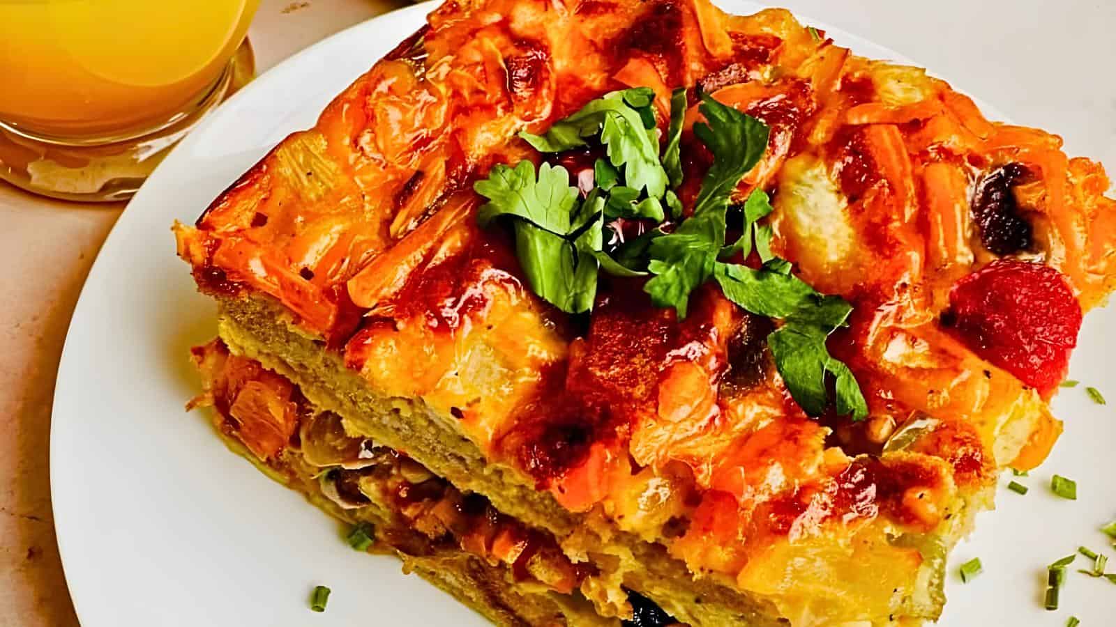 A slice of vegetable quiche garnished with parsley on a white plate, next to a glass of orange juice.