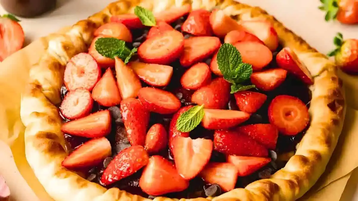 A close-up of a strawberry tart. The tart features a golden-brown crust filled with fresh, halved strawberries and garnished with a few sprigs of mint. The vibrant red strawberries contrast with the light brown crust, creating an appetizing and colorful dessert.