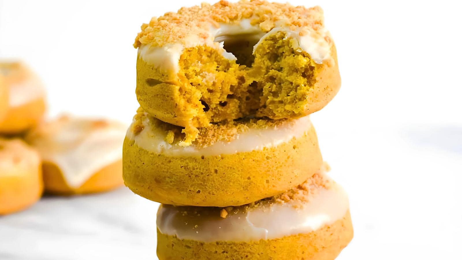 A stack of three iced pumpkin doughnuts with a bite taken out of the top doughnut, placed on a white background.