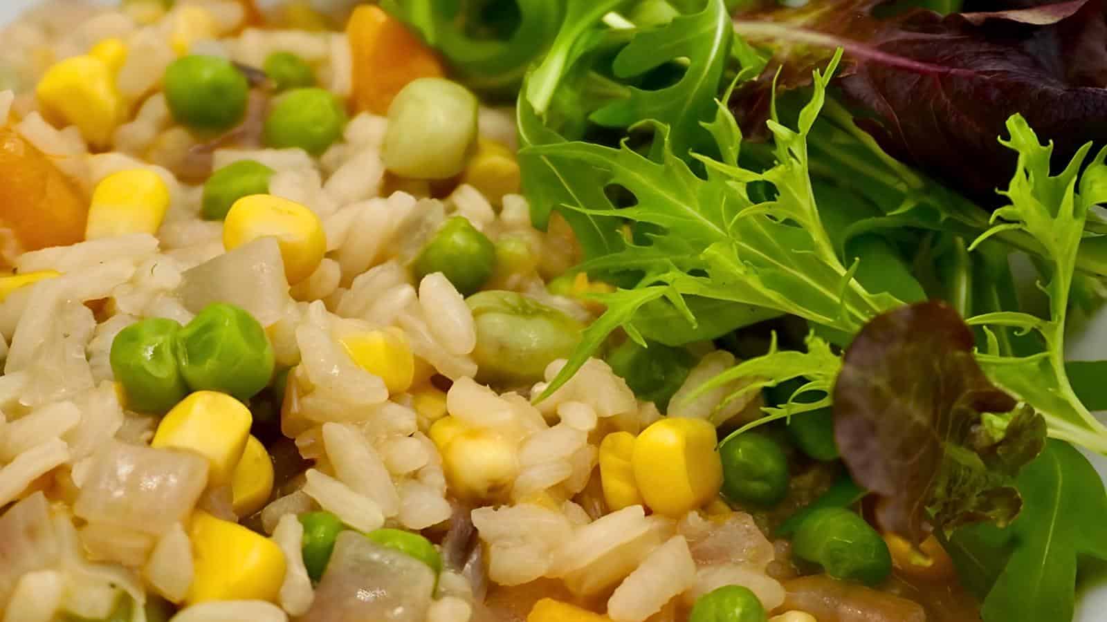A vegetable rice dish featuring peas, corn, diced carrots, and onions next to fresh green and purple salad leaves.