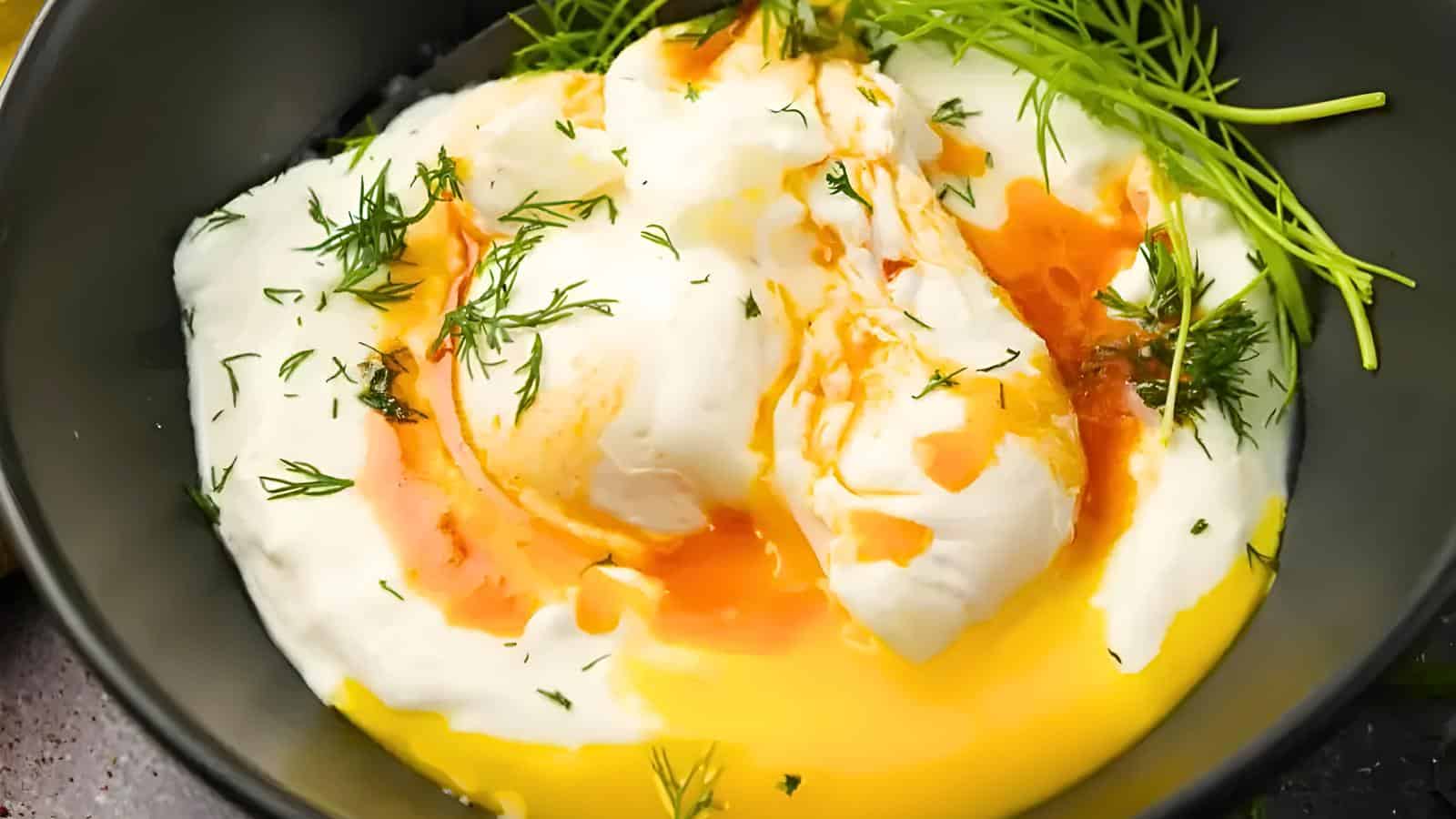 A dish featuring poached eggs topped with a drizzle of hot, melted butter and garnished with fresh dill, all served in a dark bowl.